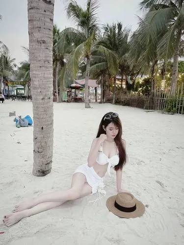 Cô gái đứng trong phòng kính đầy ánh sáng, mặc bikini trắng phối áo choàng mỏng, tay đặt lên hông, ánh sáng hắt qua lớp kính làm da trông trong và rõ nét