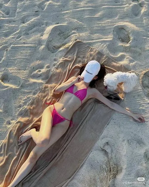 Mỹ nhân bikini hồng nằm nghiêng trên cát, tay che mặt bằng nón rộng vành trong lúc tắm nắng.