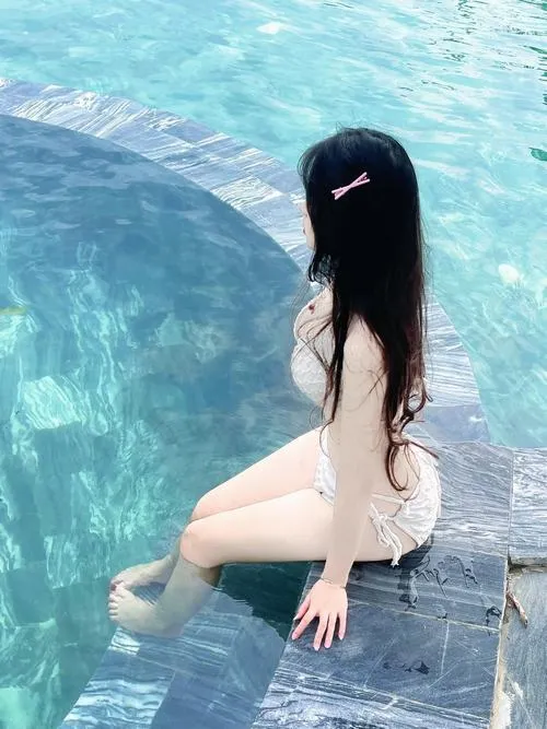 Gái đẹp mặc bikini đen đứng trên bãi cát, tay chống hông và mặt quay về phía sóng biển.