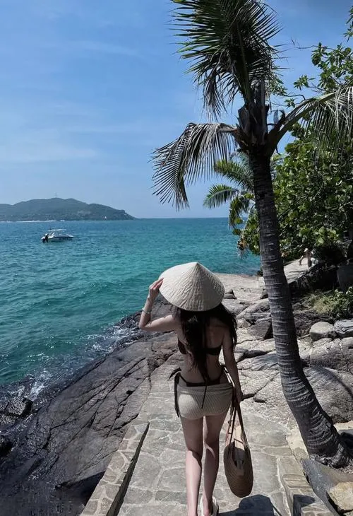 Từ góc chụp cao, cô nàng bước xuống lối đá cạnh biển trong, mũ rộng vành cùng váy trắng mỏng khiến khung hình mát mắt và khá nữ tính