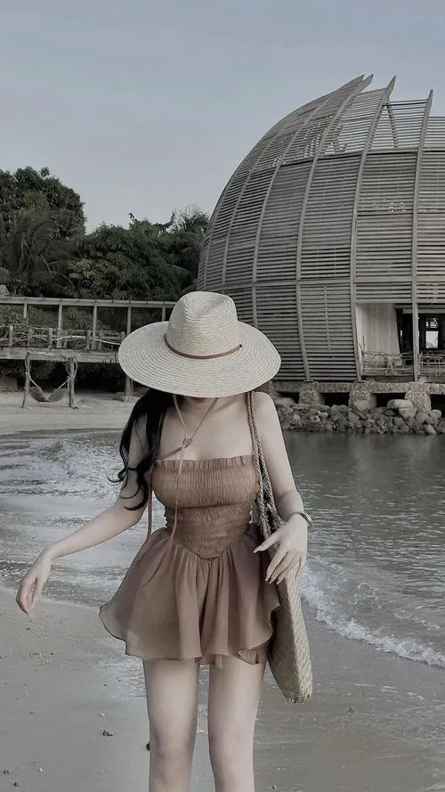Người đẹp bước ngang khu nghỉ ven biển với váy voan nâu và mũ rộng vành, tấm lưng thoáng nhẹ cùng túi xách mây làm ảnh trông rất có gu
