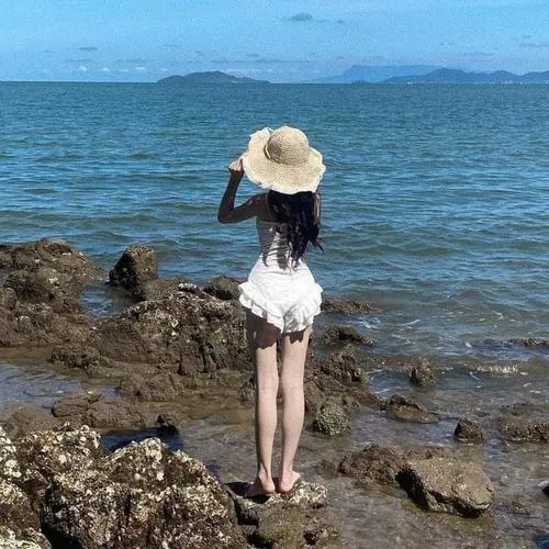 Mỹ nhân đứng trên bãi đá lổn nhổn, tay giữ vành mũ trước gió, áo trắng bèo nhẹ kết hợp chân váy tối làm người mẫu bikini bí ẩn hiện lên rất xinh