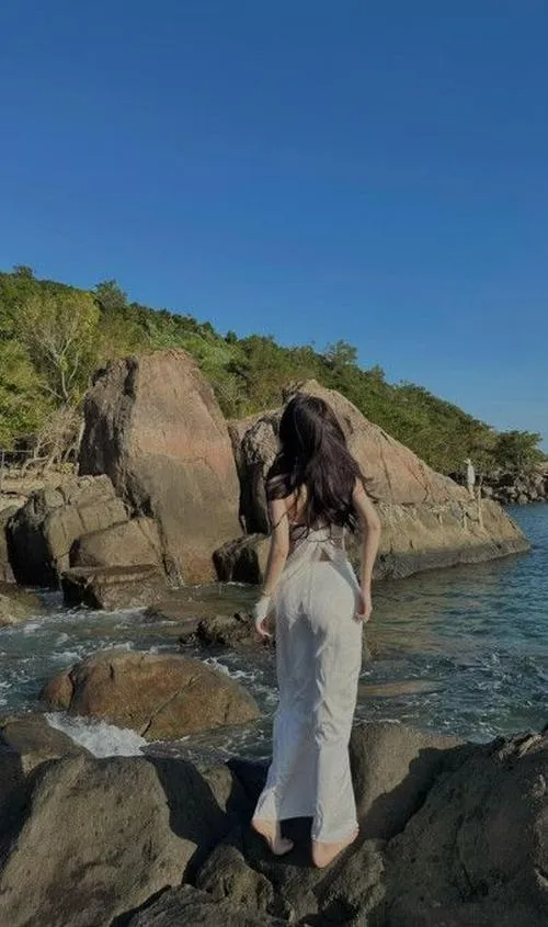 Từ xa nhìn lại, cô gái mặc váy trắng đứng trên ghềnh đá giữa vịnh nhỏ, dáng người mảnh và khoảng trời rộng gợi một vẻ đẹp kín đáo bên bờ đại dương