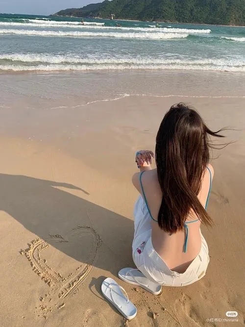 Người đẹp ngồi quay lưng trên nền cát mịn, tóc nâu rơi dài xuống lưng, đôi dép để cạnh chân làm cảnh chiều biển trông hiền và ấm