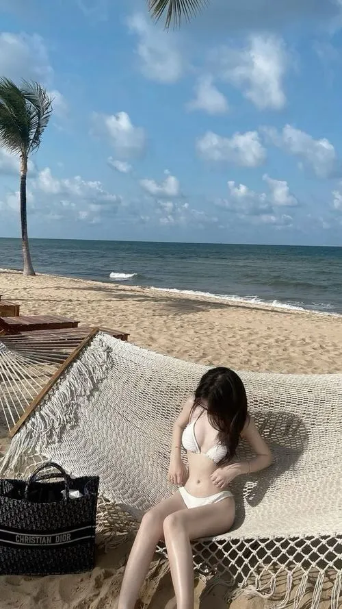 Cô gái ngồi tựa trên ghế lưới sát bãi cát, tay chống nhẹ phía sau, tóc xõa qua vai và bộ đồ sáng màu gợi đúng vibe nghỉ dưỡng rất mềm