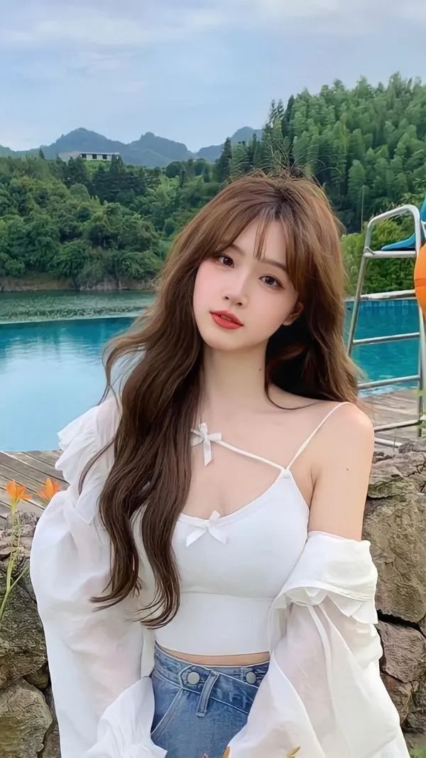 Thiếu nữ mặc áo quây trắng đứng trước dãy núi xanh, tóc nâu thả dài và ánh mắt hướng về máy ảnh làm khung cảnh ngoài trời thêm thơ mộng