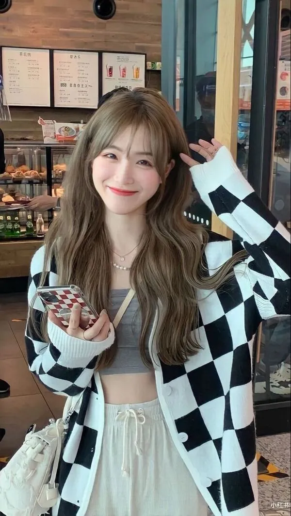 Cô nàng nghiêng người trước cửa quán, khoác cardigan caro đen trắng ngoài váy xanh nhạt, mỉm cười tươi làm khung hình trông rất xinh và sáng