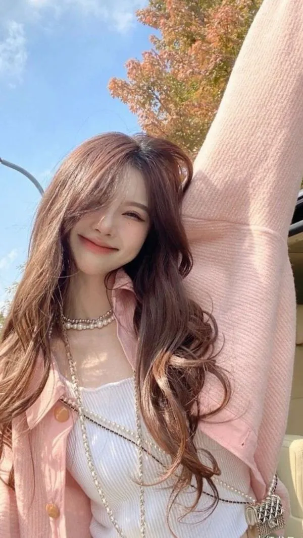 Gái đẹp giơ cao một tay giữa trời xanh, áo hồng phấn và tóc uốn lơi làm nụ cười nhẹ của cô trông ngọt ngào như ảnh lưu niệm chiều muộn