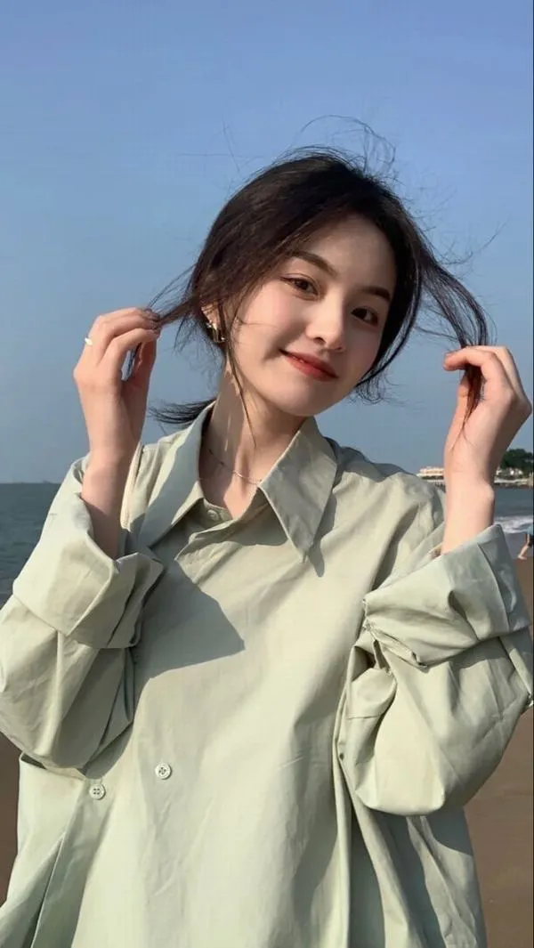 Một thiếu nữ diện đồ thể thao trắng đứng tựa hàng rào sân tennis, nâng vợt lên vai và cười lệch môi trông lanh lợi bất ngờ