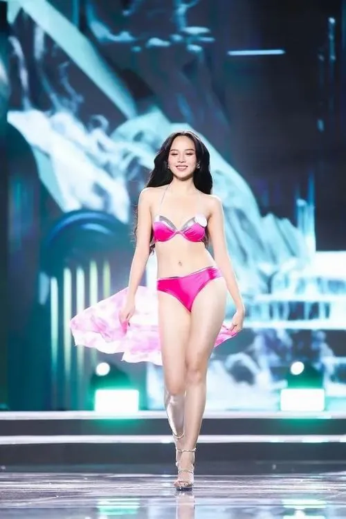 Người mẫu catwalk trong bikini hồng trên sân khấu tối, khăn voan nhỏ bay bên hông và ánh nhìn thẳng làm tổng thể khá sinh động