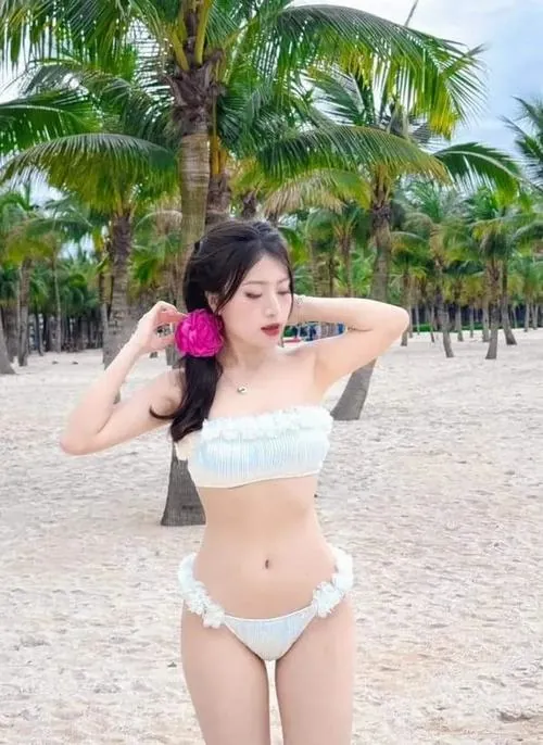Khung hình bắt khoảnh khắc cô gái chạm tay lên tóc bên hàng dừa, bikini trắng và hoa cài ngực tạo cảm giác hè rực rỡ, khá cuốn hút
