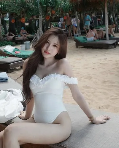 Cô nàng ngồi trên bãi cát, diện bikini một mảnh màu trắng, tay chống phía sau, ánh sáng tự nhiên tạo cảm giác nhẹ nhàng và thoải mái