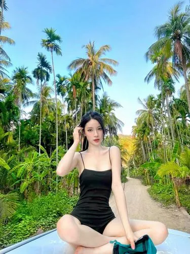 Ảnh nền thiếu nữ đứng giữa hàng dừa, khoác monokini đen đơn giản, hai tay buông tự nhiên, nền trời xanh làm nổi bật dáng người gọn gàng