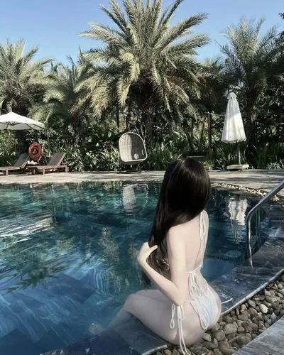 Cô gái bước chậm bên hồ bơi, tà nước xanh phản chiếu ánh sáng lên làn da, áo tắm sắc trắng làm nổi rõ phần eo và dáng đi mềm mại