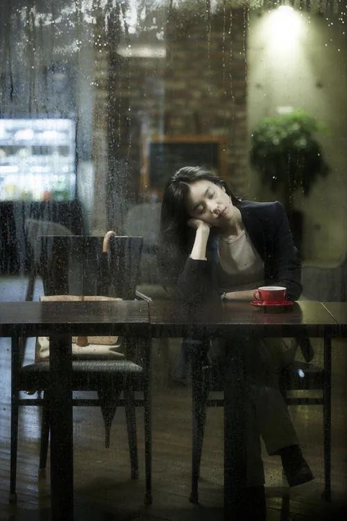 Tải hình đại diện nữ cô gái trong quán ngồi tựa đầu bên ly nước, lớp kính phản chiếu ánh đèn xanh làm khung hình mang vẻ sầu muộn