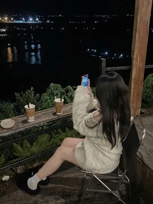 Thiếu nữ mặc áo dệt kim màu be đứng quay lưng bên quầy kính tiệm bánh đêm tay đặt nhẹ lên mép tủ và ánh đèn làm khung ảnh rất êm