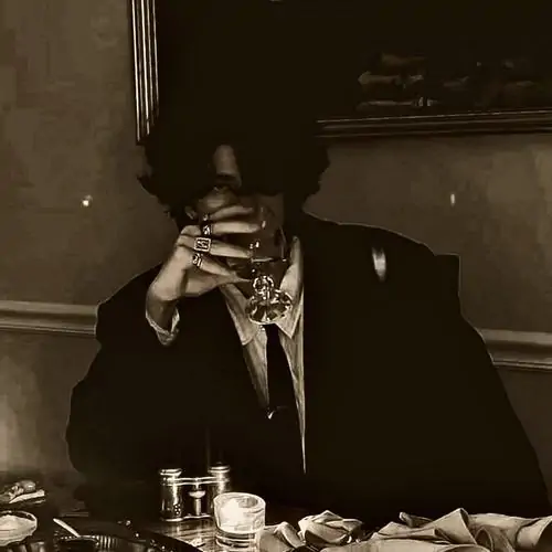 Một chàng trai ngồi bên bàn ăn tối màu, tay che mặt dưới ánh đèn vàng nâu, khói và ly thủy tinh làm không khí hơi lạnh mà cuốn