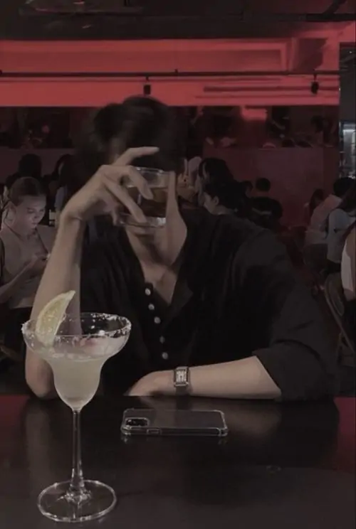 Cậu bạn ngồi trong quán ánh đỏ, tay giữ ly rồi nghiêng đầu dựa nhẹ lên vai, khung hình hơi mờ làm ảnh đẹp theo kiểu ảnh profile nam bí ẩn