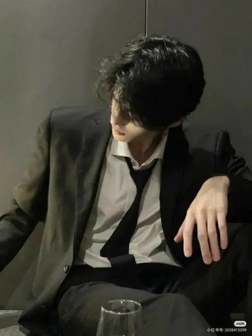 Nam thanh niên ngồi trên ghế bọc da, tay chống đầu và mắt nhìn xuống, bộ đồ tây xám rũ nhẹ khiến ảnh trông trầm và khá chỉnh chu