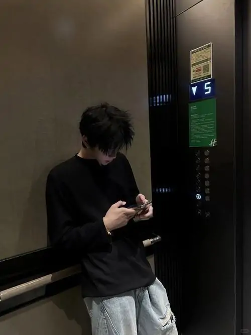 Avt Zalo cậu bạn đứng trước thang máy, đầu cúi nhìn điện thoại, áo đen và quần sáng lên ảnh ngầu nhờ bố cục gọn cùng chụp trước gương