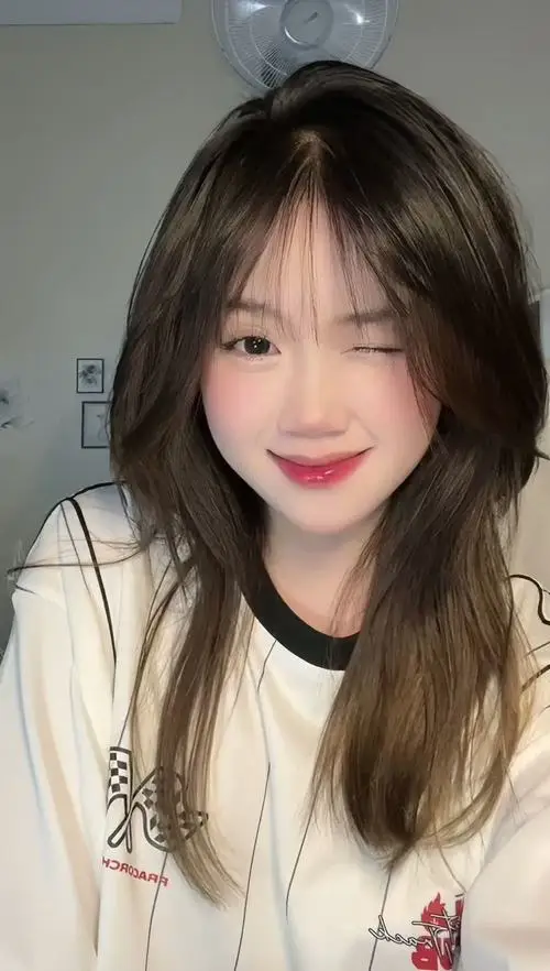 Hot girl ngồi vắt chéo chân trên ghế gỗ ngoài trời mặc quần jeans và áo len mỏng, tóc layer xinh, mắt cười cong cong rạng rỡ.