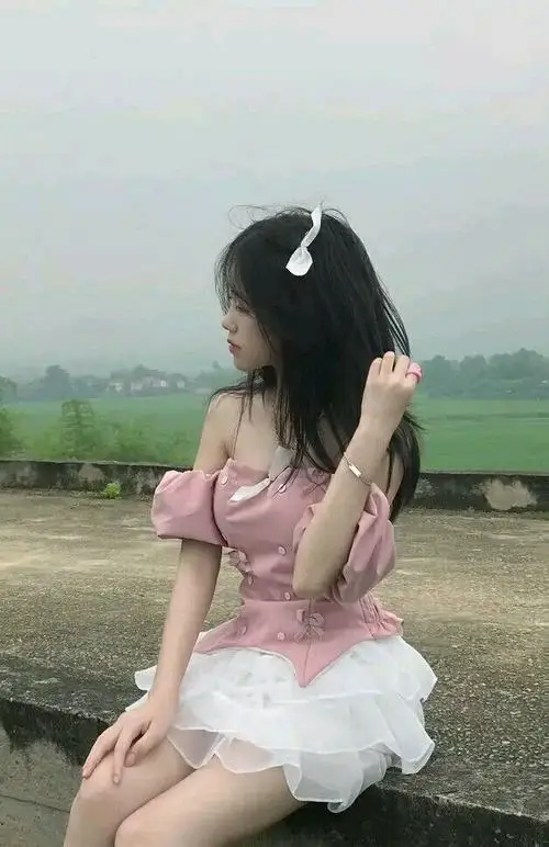 Cô đang đứng xoay nhẹ người trong phòng ngủ sáng, mặc áo thun crop và short, tóc đuôi ngựa, tay vuốt tóc với vẻ vui tươi.