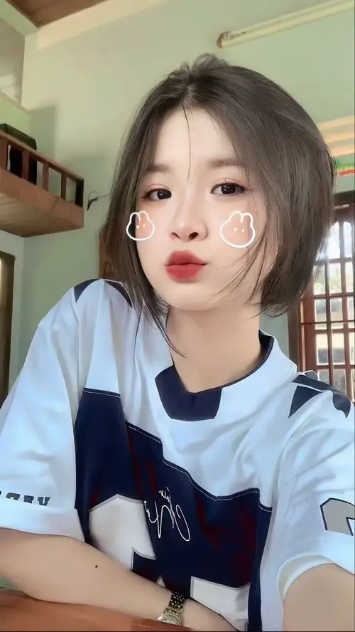 Nàng đang tựa cằm lên tay nhìn camera trong phòng sáng, váy babydoll xanh nhạt, tóc búi thấp, ánh mắt dịu dàng hồn nhiên.