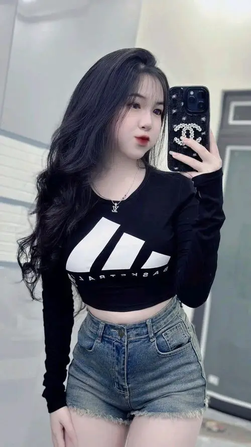 Nàng đang cầm máy ảnh nhỏ chụp ngược lại mình trong gương, váy cotton trắng, tóc layer nhẹ, môi cong cong vui vẻ trong studio sáng.