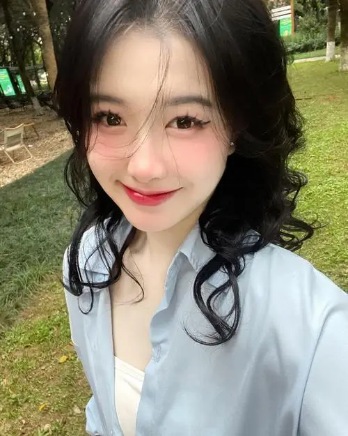 Hot girl ngồi vắt vẻo trên ghế gỗ cũ mặc quần short jeans và áo crop trắng, tay chống cằm, mắt to tròn long lanh với nụ cười rạng rỡ.