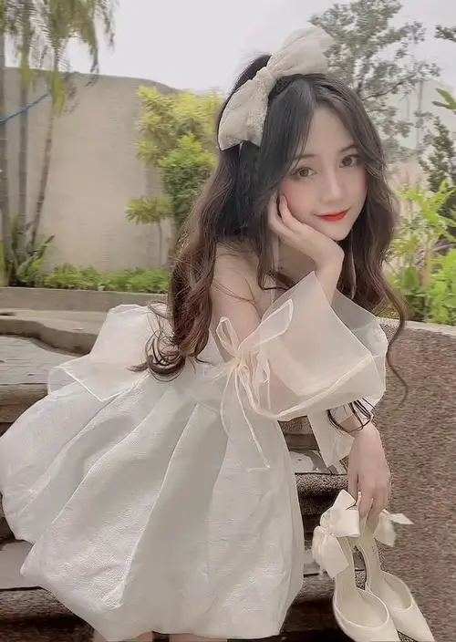 Nàng thơ đứng giữa vườn hoa nhỏ mặc váy voan dài nhẹ, tóc búi lỏng lộ cổ thanh, mắt híp lại cười duyên dáng dưới tông màu hồng nhạt dịu dàng.